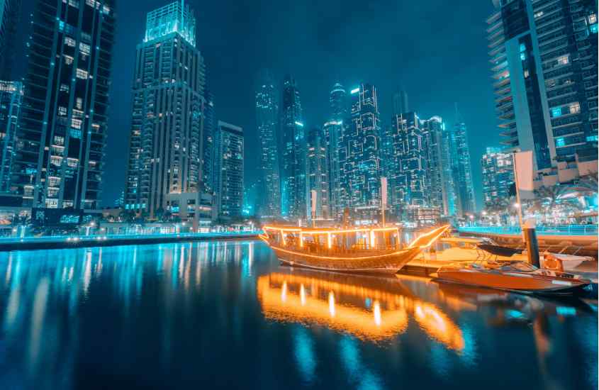 Durante a noite, barcos iluminados com luzes amarelas refletindo no mar e prédios altos iluminados com luzes brancas nos Emirados Árabes