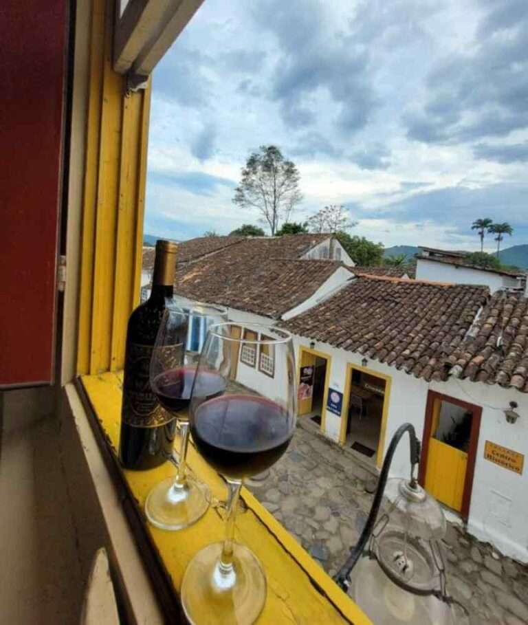 Durante um dia nublado, janela de uma pousada no centro de paraty com taças e garrafa de vinho, e rua com casas ao redor