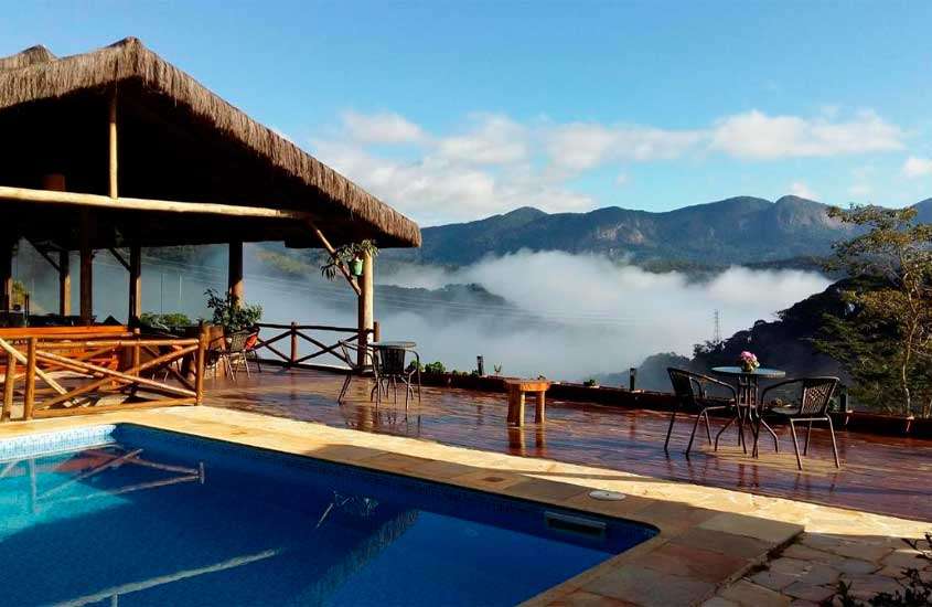 Em um dia nublado, área de lazer de um hotel em teresópolis com mesas, cadeiras, piscina, cabana de palha e horizonte das montanhas ao lado com nuvens baixas