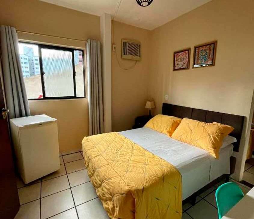 Em um quarto de pousada, cama de casal com colcha amarela, frigobar, área de trabalho, janela acortinada, quadros decorativos e ar condicionado