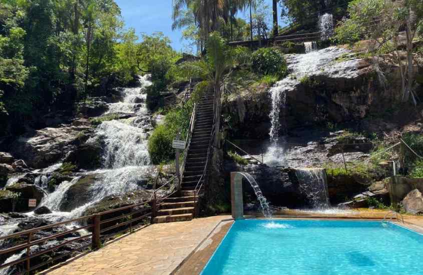 Em um dia ensolarado, área de lazer de um hotel fazenda em serra negra com piscina, cascata, cachoeira, escada de madeira e árvores ao redor