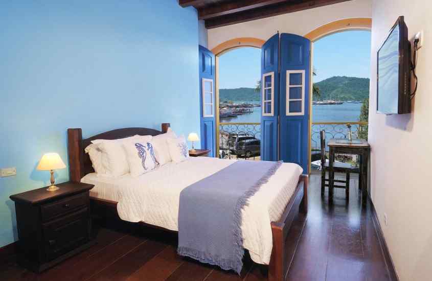 Em um dia de sol, quarto de um hotel no centro historico de paraty com cama de casal, criados e piso de madeira, janelas grandes e TV