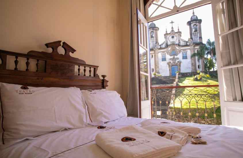 Em um dia de sol, quarto de uma das pousadas em ouro preto com cama de casal, toalhas, igreja do lado e janela grande
