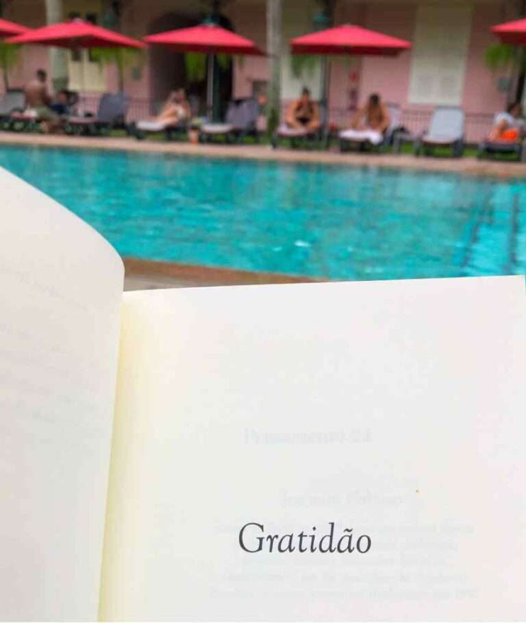 Em um dia nublado, área de lazer do hotel Vila Gale Rio de Janeiro com piscina, espreguiçadeiras, pessoas, guarda-sóis e livro escrito "gratidão"