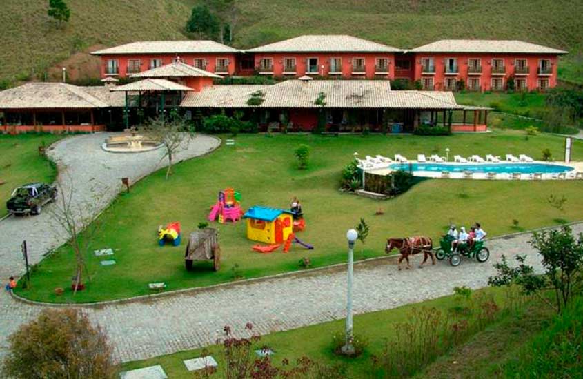 Em um dia nublado, área de lazer de um dos hotéis fazenda em teresópolis com piscina, parquinho infantil, fonte, charrete de cavalo, parte gramada com árvores