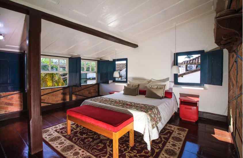 Durante o dia, quarto de horel com cama de casal, criado vermelho, estante de madeira e várias janelas ao redor