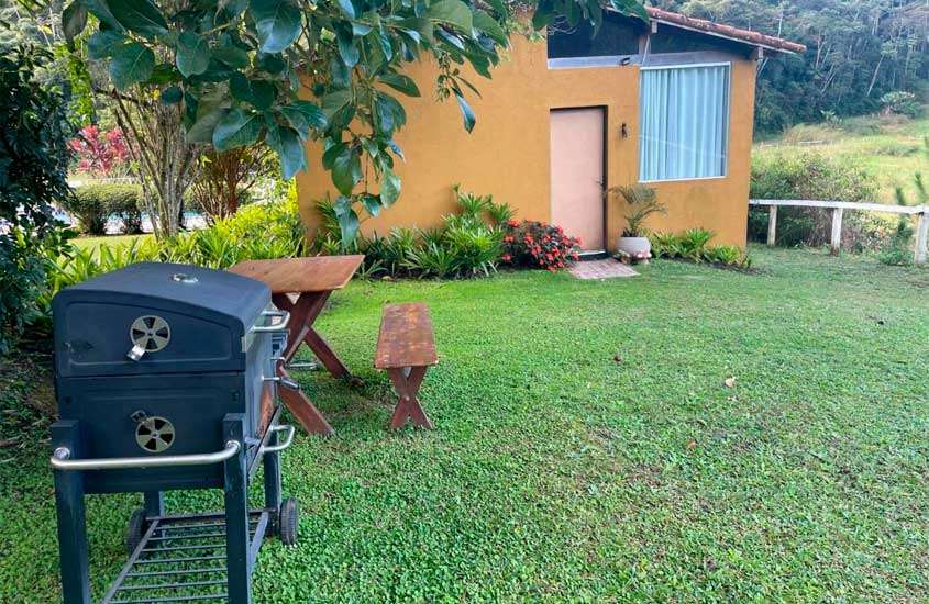 Em um dia de sol, área de lazer de um dos hotéis fazenda em teresópolis com churrasqueira, banco e mesa de madeira, parte gramada, plantas e árvores ao redor