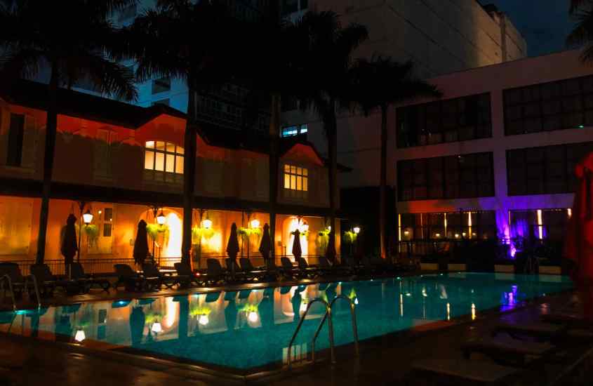 Durante o anoitecer, área de lazer com piscina, guarda-sóis, espreguiçadeiras e hotel atrás iluminando por luzes coloridas