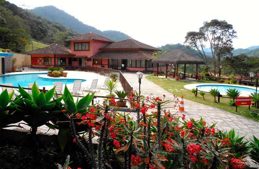 Em um dia nublado, área de lazer de um hotel fazenda em teresópolis com piscinas, plantas ao redor, espreguiçadeiras, árvores e cabana de palha com mesas e cadeiras
