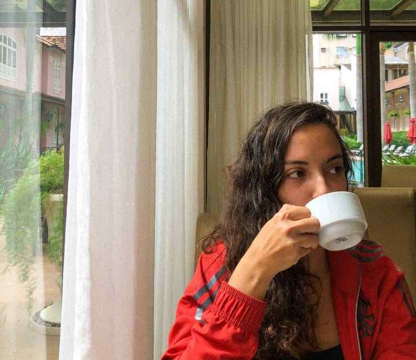 Em um dia nublado, pessoa sentada tomando café ao lado de janela grande acortinada com área de lazer do hotel atrás