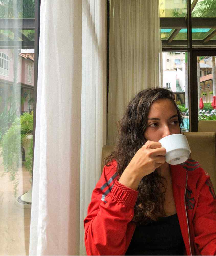 Em um dia nublado, pessoa sentada tomando café ao lado de janela grande acortinada com área de lazer do hotel atrás