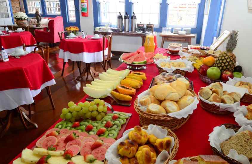 Mesa de café da manhã com frutas, pães, sucos, chás, cafpes, geleias entre outros e mesas com cadeiras ao lado
