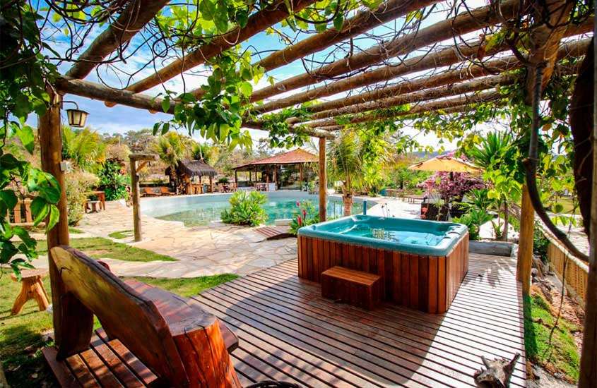 Durante um dia de sol, área de lazer de um dos hotéis fazenda próximo a brasilia com banheira, deck de madeira, banco, piscina, pergolado, parte gramada, flores e árvores decorativas