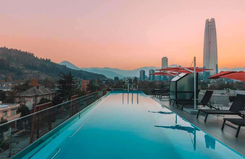 Durante o pôr do sol, área de lazer de um hotel com piscina, espreguiçadeiras, guarda-sóis, árvores e montanhas atrás