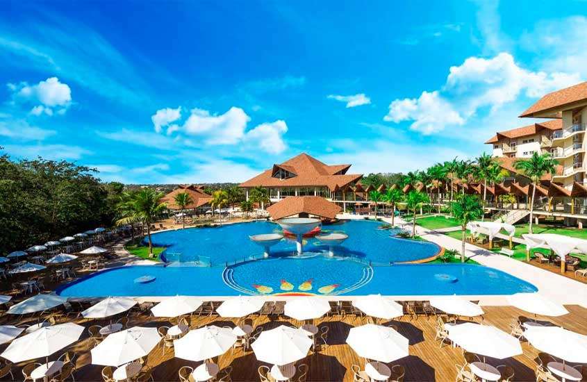 Em um dia ensolarado, área de lazer de um resort para passar o carnaval com piscina, mesas, cadeiras, espreguiçadeiras, árvores ao redor e tendas do lado