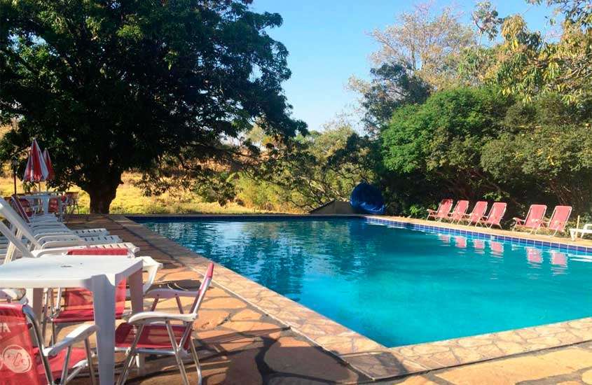 Em um dia de sol, área de lazer de um dos hotéis fazenda próximo a brasilia com mesas, cadeiras, guarda-sóis, espreguiçadeiras, piscina, árvores e parte gramada