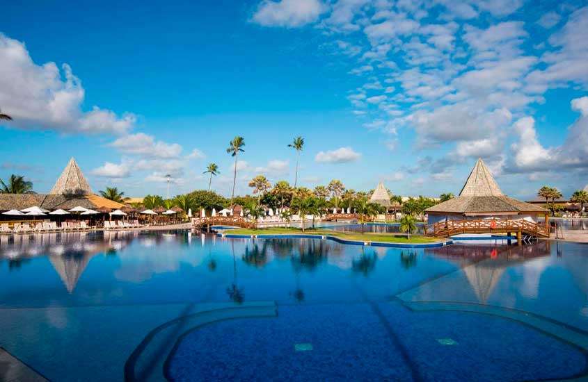 Em um dia de sol, área de lazer de resort para passar o carnaval com piscinas, ponte de madeira, árvores ao redor, espreguiçadeiras, guarda-sóis e tendas