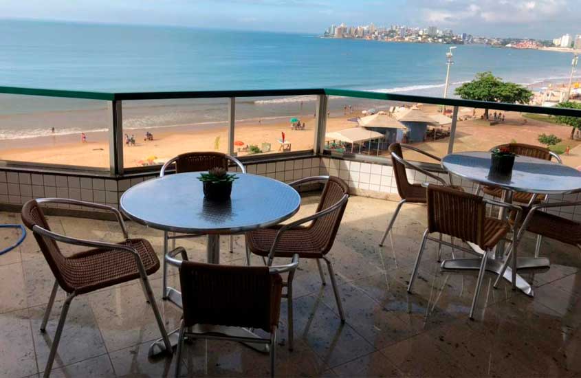 Em um dia de sol, varanda de um apartamento com vista da praia, mesas, e cadeiras