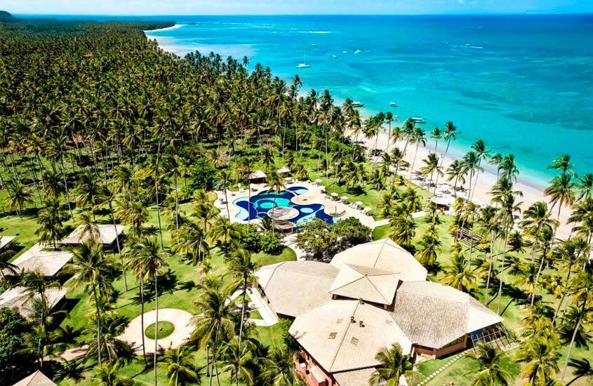 Em um dia de sol, vista aérea de resort para o carnaval com praia do lado, árvores ao redor e piscinas no meio