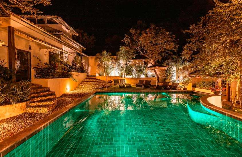 Durante a noite, área de lazer de um hotel para passar o feriado em Brasília com piscina, espreguiçadeiras, árvores e plantas decorativas ao redor