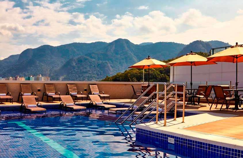 Em um dia de sol, área de lazer de um hotel com piscina, espreguiçadeiras, guarda-sóis e paisagem das montanhas