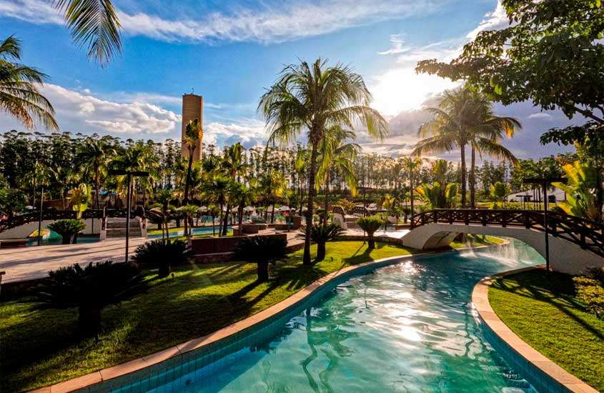 Em um dia de sol com nuvens, área de lazer de um resort para passar o carnaval com piscinas, ponte, árvores, plantas e parte gramada ao redor