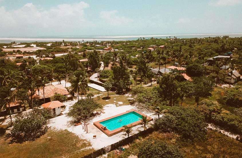 Em um dia de sol, vista aérea de um hotel onde ficar nos Lençóis Maranhenses com piscina, dunas, mar e árvores ao redor