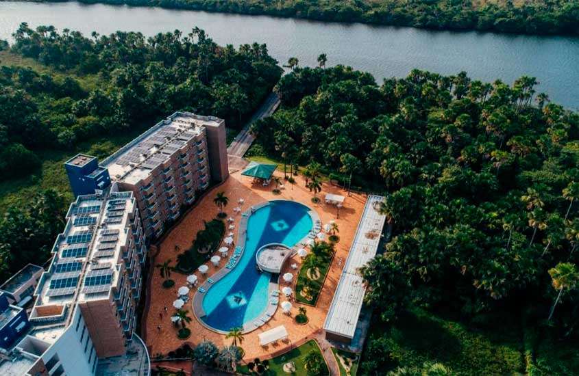 Em um dia de sol, vista aérea de hotel com piscina, árvores ao redor e rio do lado
