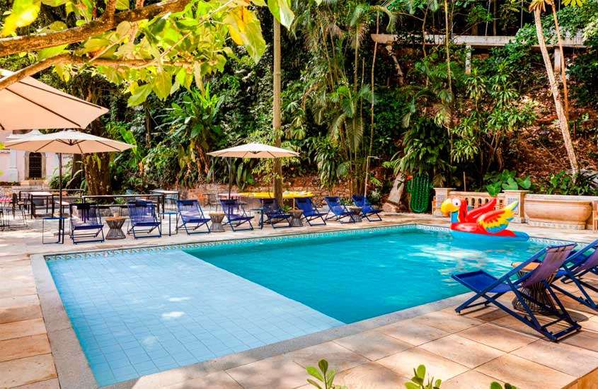 Durante uma manhã de sol, área de lazer de um dos hotéis para passar o réveillon no Rio de Janeiro com espreguiçadeiras, piscina, boias, guarda-sóis e natureza ao redor