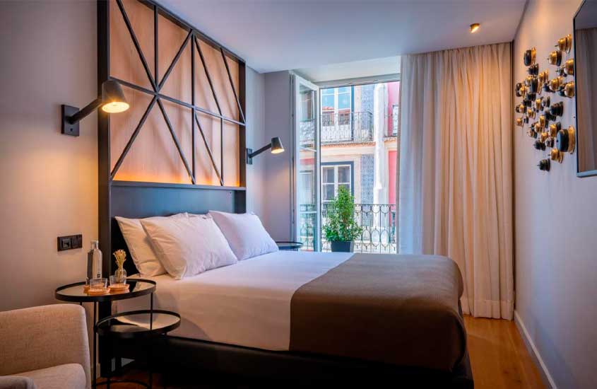 Quarto de um hotel com cama de casal, criados de metal, poltrona, TV, janela grande acortinada, luminárias e vaso de planta