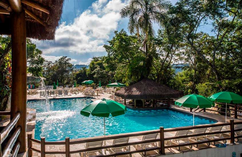 Em um dia de sol, área de lazer de um hotel com piscina, bar molhado, espreguiçadeiras, cascata, guarda-sóis e árvores ao redor