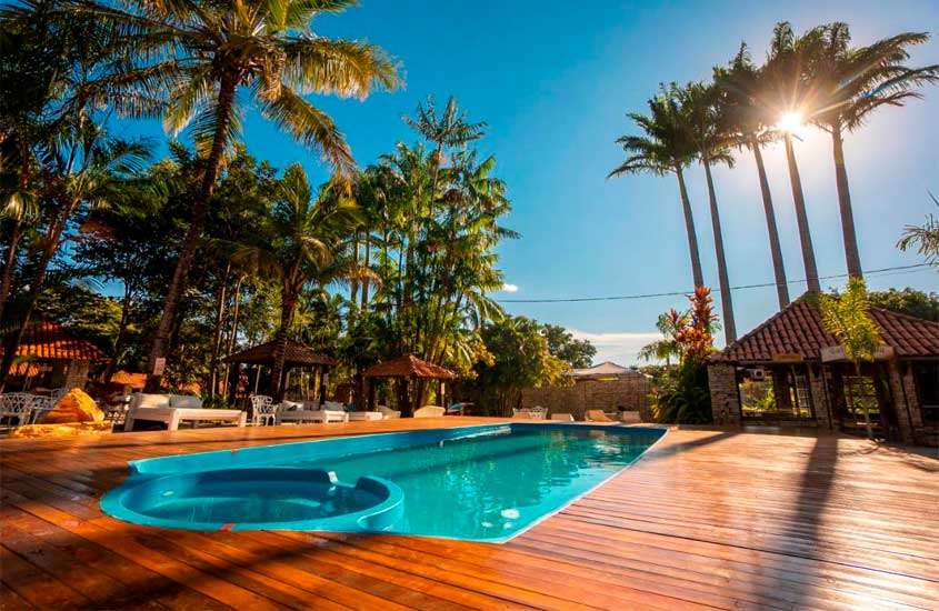 Em um dia ensolarado, área de lazer de um dos lugares perto de Brasília para passar o feriado com piscina, deck de madeira, cama, mesas, cadeiras, espreguiçadeiras e árvores ao redor