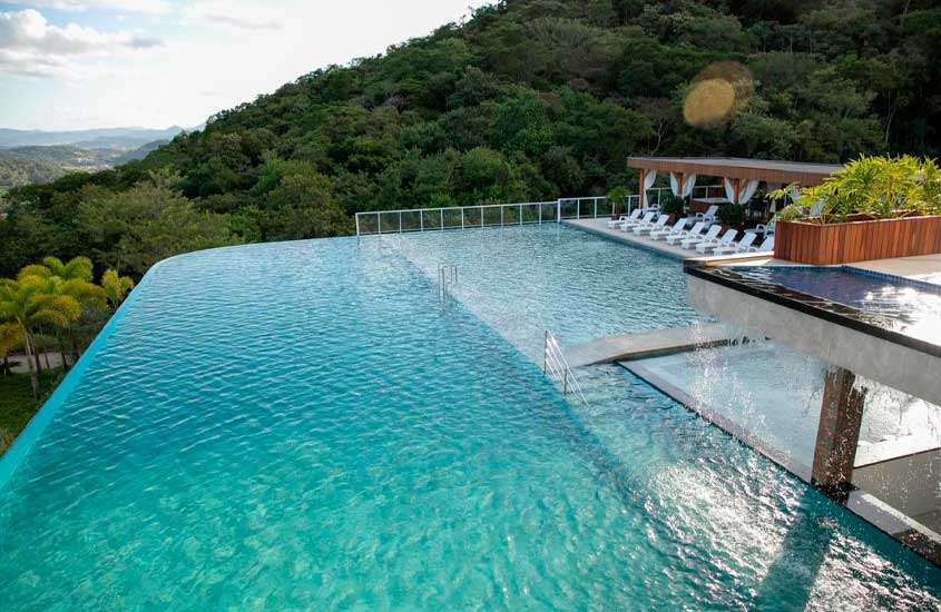 Em um dia de sol, área de lazer de um resort com piscinas grandes de borda infinita, espreguiçadeiras, plantas ao redor e reserva do lado