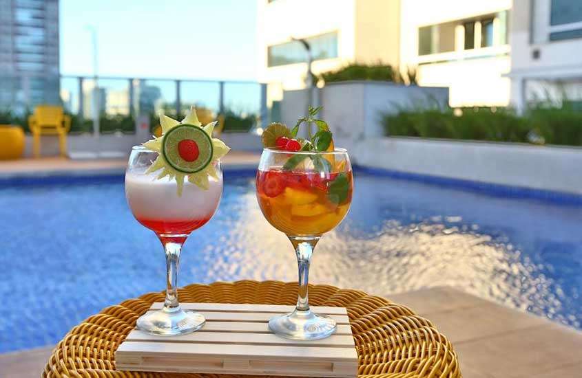 Em um dia de sol, área de lazer de um hotel com piscina, cadeira, plantas ao redor e coquetéis de frutas