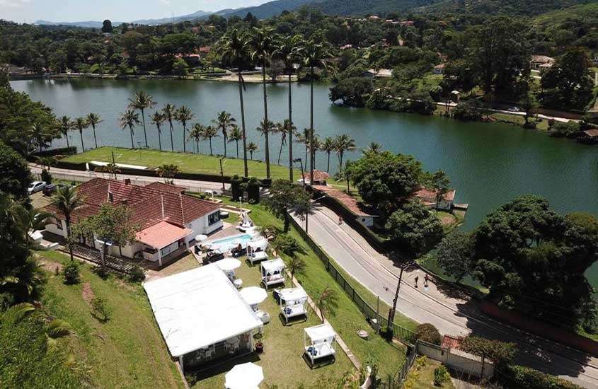 Durante o dia, vista aérea de um hotel com piscina, área de descanso, lago do lado e árvores ao redor