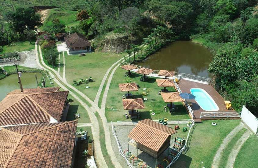 Em um dia de sol, área de lazer de uma pousada com piscina, lago, tendas, playground, árvores e parte verde ao redor