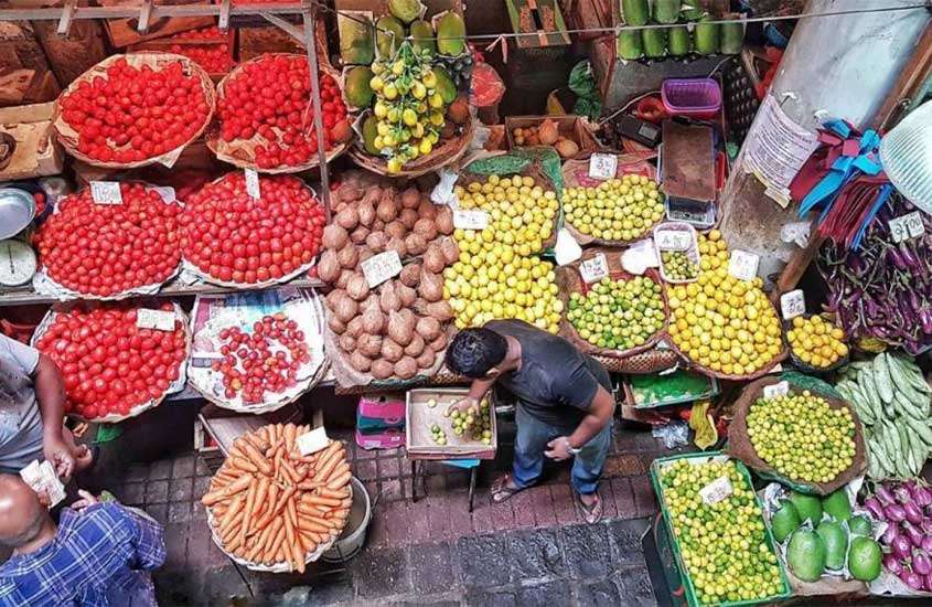 Barraca de mercado com diversas frutas coloridas, com vendedores e clientes ao redor
