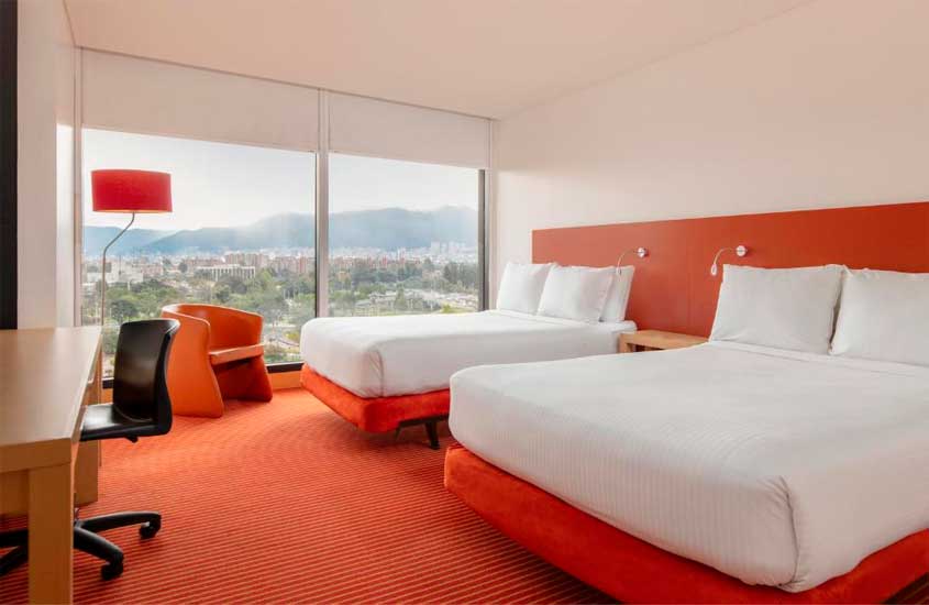 Quarto de um hotel onde ficar em bogotá com camas de casal, área de trabalho, luminária, poltrona e janela grande acortinada