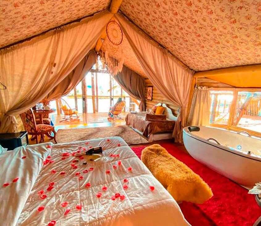 Quarto de um dos hotéis fazenda na serra gaúcha cama de casal com pétalas de rosa, tapetes, banheira de hidromassagem, mesas, cadeiras, sofá, cortinas e varanda com cadeiras de balanço