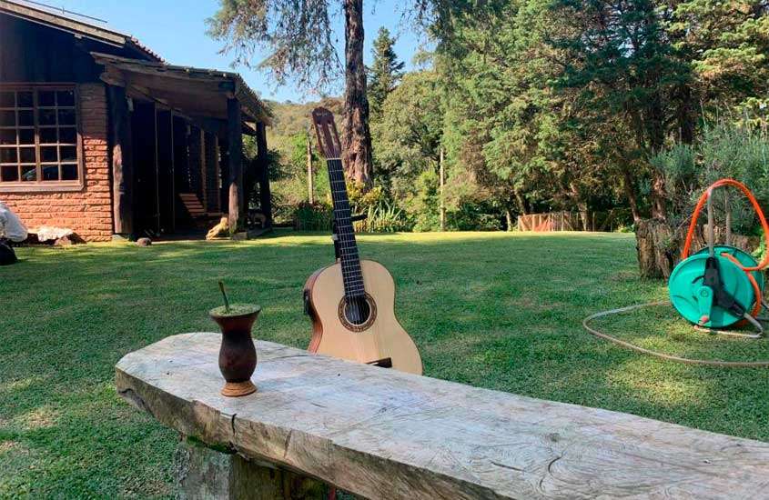 Em um dia de sol, área lazer de um hotel fazenda na serra gaúcha com violão, banco de madeira, árvores e plantas ao redor