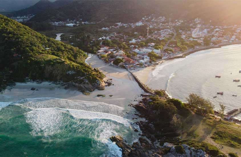 Em um final de tarde, paisagem aérea de praia em Florianópolis, lugar onde ir no feriado de corpus christi, com praia, árvores ao redor e cidade no fundo