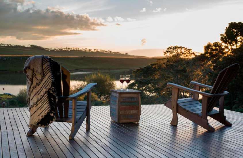 Durante o final da tarde, deck de hotel fazenda em santa catarina com cadeiras de madeira, mesinha, taças de vinho e paisagem de lago com árvores ao redor