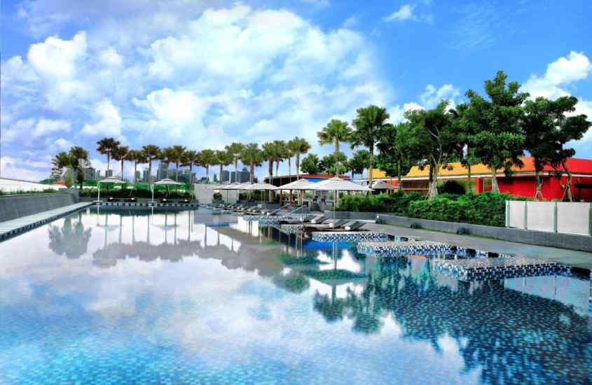 Em um dia de sol, área de lazer de um hotel em singapura com piscina, espreguiçadeiras, guarda-sóis e árvores ao redor