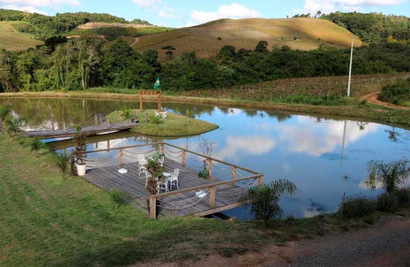 Em um final de tarde, lago de hotel fazenda na serra gaúcha com deck de madeira, ponte pra ilha, árvores e serras ao redor