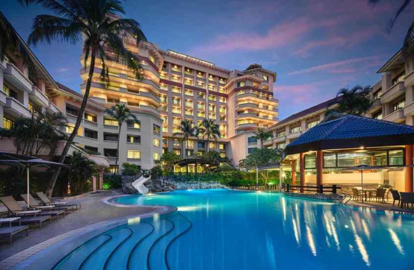 Durante o entardecer, área de lazer de um hotel em singapura com piscina, mesas, cadeira, guarda-sóis, espreguiçadeiras, árvores e luzes ao redor