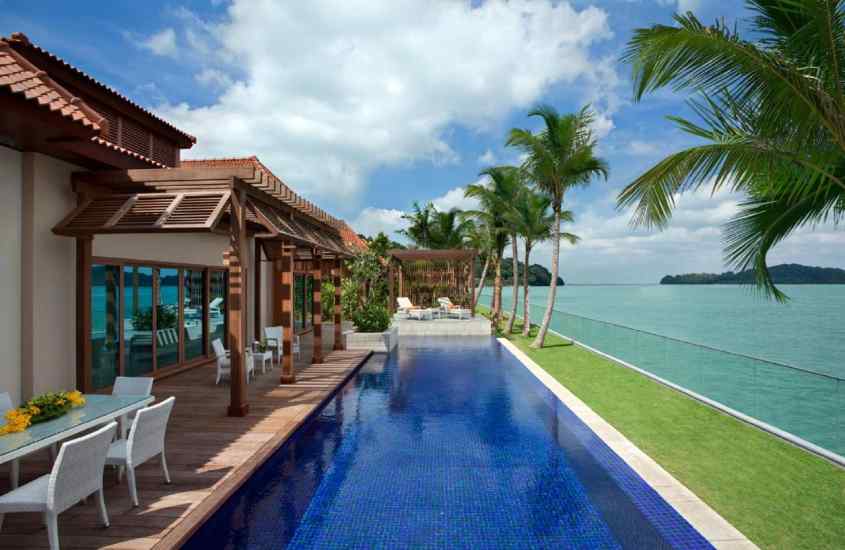 Em um dia de sol, área de lazer de um hotel em singapura com piscina, mesa, cadeiras, camas, árvores e mar ao lado