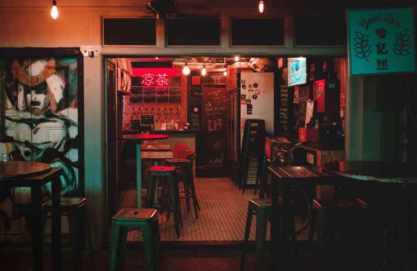 Durante a noite, restaurante de um bairro onde ficar em singapura com mesas, bancos, luminária vermelha, TV, geladeira e caixa