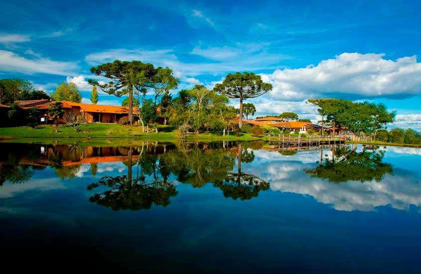 Em um dia de sol com nuvens, lago de hotel fazenda em santa catarina com árvores ao redor ponte e hotel atrás