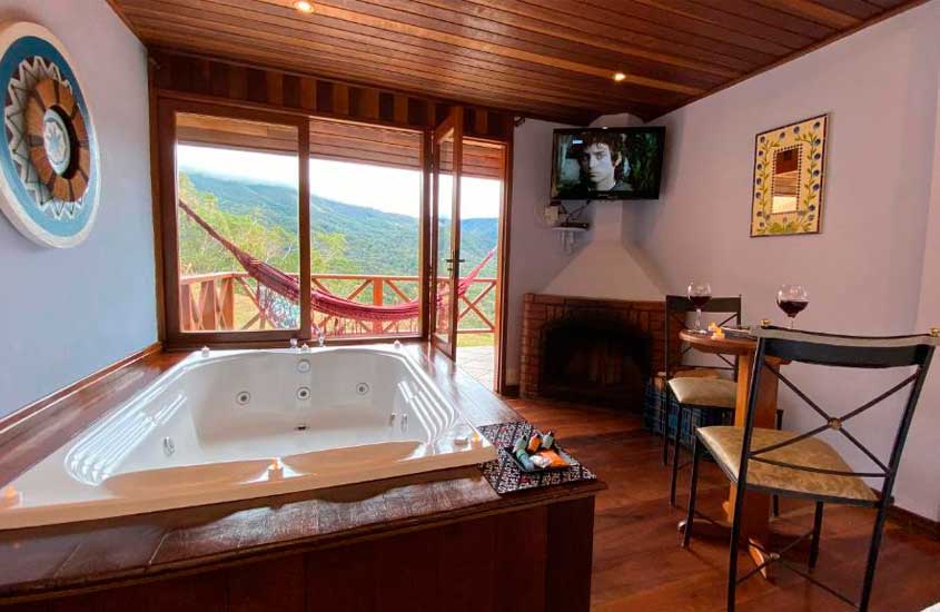 Área de banheira de um dos lugares românticos para viajar no rio de janeiro com lareira, mesa, cadeiras, taças de vinho, quadros decorativos, TV e rede na varanda