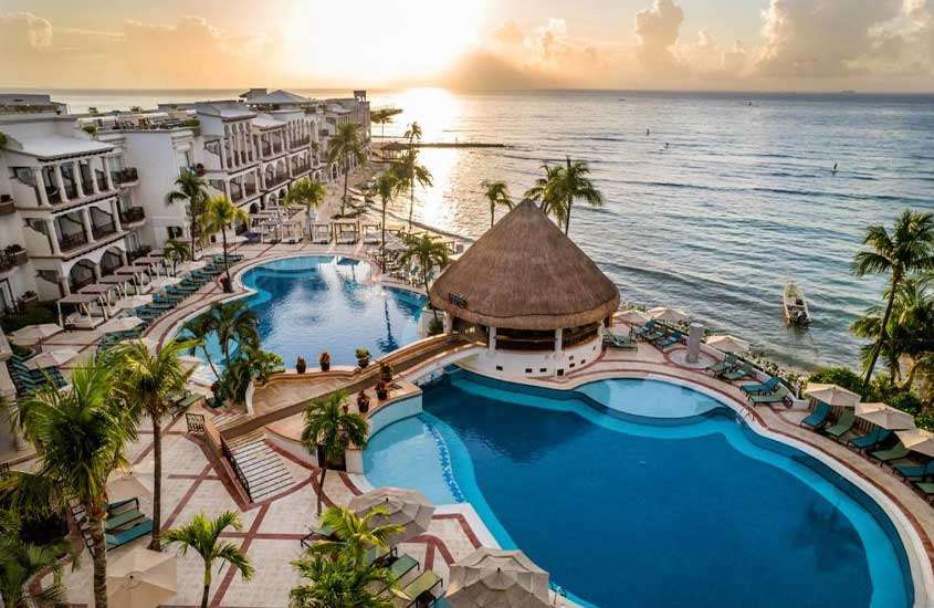 Durante pôr do sol, vista aérea de hotel onde ficar em Playa del Carmen com mesas, cadeiras, espreguiçadeiras, piscinas, tendas, guarda-sóis, bar no mar e árvores ao redor